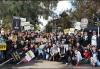A very large, diverse group outside in a park setting. Those in the front two rows either sit or kneel on the concrete. Visible throughout are black and white SAG-AFTRA picket signs; there is also a cardboard sign with "Union" in large, all-cap copy.