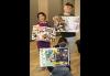 Clockwise from left, Yamashita (Asian), Sparks and Schwartz, kneeling, respectively hold up a sheet of paper. Each sheet has magazine cutouts and photos. Schwarz has on a protective face mask.