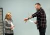 Jacques, left, and Adams face one another while holding sides. Jacques wears a gray geometric sweater and pants. Adams wears a flannel shirt and dark jeans; his tattoo is visible. Adams's left arm is outstretched and he is captured mid-speech.