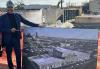 Knight (Black), outside at an active construction site. He is dressed in a dark suit and wears a hard hat. With his left hand, he holds up a very large photo of East End Studios when once its completed; he points to the photo with his right.