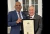 Knight (Black) and DiNapoli hold up a framed certificate. Knight is in a blue blazer and light blue dress shirt; DiNapoli is in a dark-brown blazer, a white dress shirt and tie.