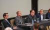 A wide shot of four male panelists, including Crabtree-Ireland, second from right, seated at a table. Crabtree-Ireland is speaking. Matz and Rushing are on the far left; Doroshow, far right, turns to face Crabtree-Ireland.