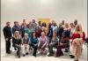 A photo of a large, diverse group of people in a nondescript room. They are arranged in three or four rows with those in front seated. Behind the group is a standing SAG-AFTRA banner.