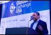 Rodriguez stands behind a large podium with a microphone. He wears a dark blue blazer over a light blue dress shirt; an earpiece with a microphone is in his ear. He is captured mid-speech. Behind him is a large banner bearing the Global IP Exchange USA logo and various event sponsors.