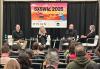 In the background, four people are seated onstage in chairs. The second person is captured mid-speech as the others listen on. Behind them is a SXSW 2025 poster. In the foreground is the audience whose backs are to the camera.