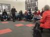 Kehoe stands in front of a group of people seated in chairs arranged in a semicircle. Her back is toward the camera.