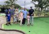 SAG-AFTRA Hawaii Local young performers, their parents and friends stop for a quick picture at the first hole before playing the “Pali” course at Bayview Mini Golf. A diverse group of children and their parents stand smiling at a mini golf hole. The green area by the hole is made of artificial turf and a concrete path with a gray brick boundary lies adjacent to the green and hole. There are also tropical trees.