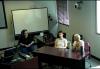 Three individuals are in a classroom setting. Two sit behind a table while one on the far left is in an office chair. The person on the far right is captured mid-speech while the other two listen intently.