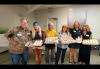 Six individuals are in a lobby-like setting. Four people hold trays of cupcakes; one person, second from the right, holds a small cake.