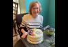 An individual is seated at a table. She wears glasses and a striped sweater. In front of her is a cake with white frosting, colorful flowers and “Happy Retirement Jayne!” written in purple frosting.