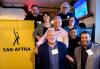 A smiling group of diverse individuals gather next to a yellow standing SAG-AFTRA banner in a restaurant setting.
