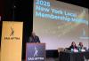 Knight stands on a podium with SAG-AFTRA on a black background in white text. To his left is a yellow banner with SAG-AFTRA in black text. There is a large screen behind him with the “2025 New York Local Membership Meeting” title card. Two individuals sit to his right behind a table.