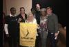 A diverse group of men and women stand for a photo against a gray wall. A women, center, holds up a small yelllow banner bearing the SAG-AFTRA logo. A diverse group of men and women stand for a photo against a gray wall. A women, center, holds up a small yelllow banner bearing the SAG-AFTRA logo.