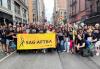 SAG-AFTRA members gather before the Pride march begins. A large group of people gather behind a yellow parade banner with a black SAG-AFTRA logo in the center. The people wear black T-shirts with a rainbow and white SAG-AFTRA logo in the center. Some hold teal fans with a rainbow and white SAG-AFTRA logo.