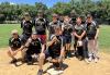 A group of 13 softball players pose on a softball field. The players are wearing black jerseys with a yellow SAG-AFTRA logo in the center. There are green trees and a blue sky in the background.