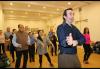 Un caballero a la derecha pantomina un movimiento de baile. Detrás de él, un grupo diverso de hombres y mujeres, sigue sus movimientos.