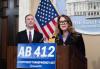 Fisher is behind a podium with a sign for AB 412. She wears a blazer over a blue shirt, jewelry and glasses. She holds the microphone’s neck as she speaks. Behind her, a person listens intently. Also visible is the American flag and an image of the California State Capitol building.