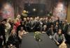 Dozens of well-dressed attendees sit and stand around a table covered in a black tablecloth.