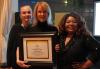 From left, Duerr, Burkhead and Underdue stand closely together. All three women wear black dresses, with Underdue’s dress sequined. Burkhead holds a framed certificate. The top and the bottom of the certificate are boarded with a yellow line; the text is illegible.