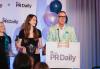 At a podium branded with the words PR Daily, a woman on the left wearing a black dress holds a crystal statuette engraved with the words PR Daily Social Media & Digital Awards; on the right is a man in a floral pattern suit, turquoise pocket square and black-framed glasses speaking into the podium’s microphone.