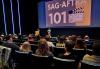 Attendees watch the SAG-AFTRA presentation at the Arizona State University MIX Center on Nov. 8. An audience seated in a theater. The setting is a dimly lit auditorium with vertical blue light strips illuminating the walls. On the stage at the front of the room, two presenters stand under a large screen displaying a slide that reads "SAG-AFTRA 101" with an image of a clapperboard. One presenter stands at a podium, while the other is speaking. The audience includes individuals of various ages, attentively facing the presenters.