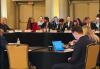 Several individuals are seated around a table in a large room. Wood Einertson, second from far left, wears a red blazer over a black shirt. They are turned to another person.
