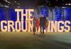 A wide shot of four individuals, three women and one man, standing outside in a grassy area. Behind them is a display of lighted letters that spell “The Groundlings” in all caps. The Downtown Los Angeles skyline is visible above a tall wall. Some blue lighting is visible.