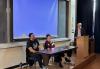 Alton and Elmaleh are seated at a small table in the front of a lecture hall. Dr. Griffey stands to the far right and looks toward the two individuals. Behind them is a projector screen that glows blue.
