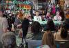 From left, moderator and National and Hawaii Local Board member Henry Ian Cusick, Natalie Alyn Lind, Kai Caster, Layne and Miles Ulrich. Five people sit at a table in folding chairs, smiling, on a set made up to look like a halloween store. In the foreground can be seen the backs of audience members.