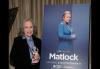 Kathy Bates, miembro de SAG-AFTRA, celebra recibir el Premio a la Persona de Televisión del Año en la 62.ª edición de los Premios ICG a los Publicistas. Foto: Evans Vestal Ward. Bates junto a un póster del programa de televisión "Matlock" en una habitación alfombrada con poca luz. Bates viste un traje pantalón negro y una camisa azul, y sostiene su estatuilla de premio. El póster está apoyado sobre un gran caballete.