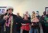 In the foreground, a man impersonating Elvis Presley is singing with his left arm extended. He wears a black jacket, a leopard-print scarf and pink lei, gold-framed sunglasses, sideburns and a Santa Claus hat. Behind him in two rows are a group of singers. Many wear holiday festive clothing: red or green and Santa Claus hats.
