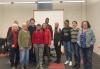 Twelve people take a photo together in a classroom-like environment. They are arranged in uneven rows, with a greater grouping on the left side of the photo. Behind them is a whiteboard.