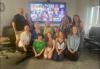 A group of people inside a darkened room. The group frames a flatscreen television with those in front kneeling on the carpet and those on the side standing. The flatscreen displays a Zoom meeting with other individuals.
