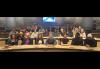 Un grupo grande y diverso está sentado en filas de mesas largas dentro de un auditorio o escenario similar a un teatro.