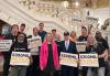 A group of diverse individuals picket inside a government building with a large white staircase. Many in the photo hold protest signs with the text, "$300MIL PA Film" with "$300MIL" in dark color and "PA Film" in yellow.