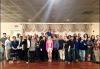 A very large, diverse group takes a photo in a ballroom setting. Aside from a woman dressed in pink, center, others in the photo are dressed in dark colors.