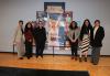 Six individuals stand in front of a painted collage. They are grouped in two sets of three and there is a large gap between them. Visible is a painting of a person holding a sign that reads "Union" in all-cap lettering.