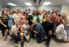 A diverse group of casually dressed people pose together in a classrom