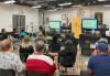Film Louisiana President Jason Waggenspack speaks to SAG-AFTRA members in New Orleans. A speaker stands at the front of a spacious well-lit room, gesturing while addressing a diverse group of people seated in rows. Behind the speaker, a screen displays "Film Louisiana" alongside a yellow SAG-AFTRA banner. People seated in chairs listen.