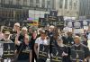 SAG-AFTRA members gather with New York Local Board members Ray DeForest and Marc Baron; Chief Labor Policy Officer & New York Local Executive Director Rebecca Damon; Executive Vice President Linda Powell; and Vice President, New York and New York Local President Ezra Knight to show their solidarity with the American Federation of Musicians. A group of people are holding black signs with the white SAG-AFTRA logo, reading “SAG-AFTRA in Solidarity” in white and yellow text. There are people to the right of the image with navy and white signs that read “Musicians United: Better Healthcare.”