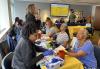 SAG-AFTRA members receive free health screenings at the SAG-AFTRA N.Y. Health Care Safety Net Committee Health Fair. A group of people sit on two sides of a table covered with a yellow tablecloth. The people on the left side perform finger sticks and blood pressure readings on the people on the right side.