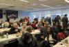 New York Local members enjoy food and refreshments at the New York local office. A large, diverse group of people seated in rows at tables inside a room. There are plates of food on the table. A yule log video appears on the screens in the background.