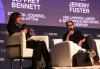 Fuster y Bennett están sentados en una silla y un sofá blancos respectivamente. Fuster está vestido de negro y sostiene un micrófono de mano; se le capta en medio de un discurso. Bennett lleva ropa negra y botas color canela; hay un maletín negro a sus pies. Él también sostiene un micrófono. Está sentado con las piernas cruzadas y el cuerpo ligeramente girado hacia Fuster. Detrás de ellos hay una pantalla de proyección con sus nombres y títulos.