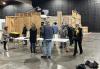Members tour Harbor Island Studios. People in casual clothing mill around a large warehouse-like space. There are plastic tables and, in one part of the room, there are free-standing wooden walls of a set.