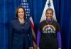 Harris and Lyman Harris, left, wearing a blue suit, stands next to Lyman, who is wearing all black, including a SAG-AFTRA shirt with the union’s logo and a rainbow. Behind them is a blue backdrop and two flags, including an American flag.